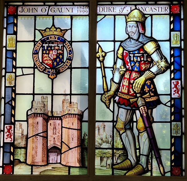 Photo of a stained glass window. There's a title across the top reading 'John O'Gaunt, 1340-1399, Duke of Lancaster'. The window is divided into two parts: To the left is an image of the substantial stone gatehouse of Lancaster Castle, beneath a coat of arms with the motto 'honi soit qui mal y pense'. To the right is a man in plate armour. His helmet is in the form of a golden crown. He has a colourful tabard covered in heraldic symbols, and his hand rests on a sheathed longsword at his side.